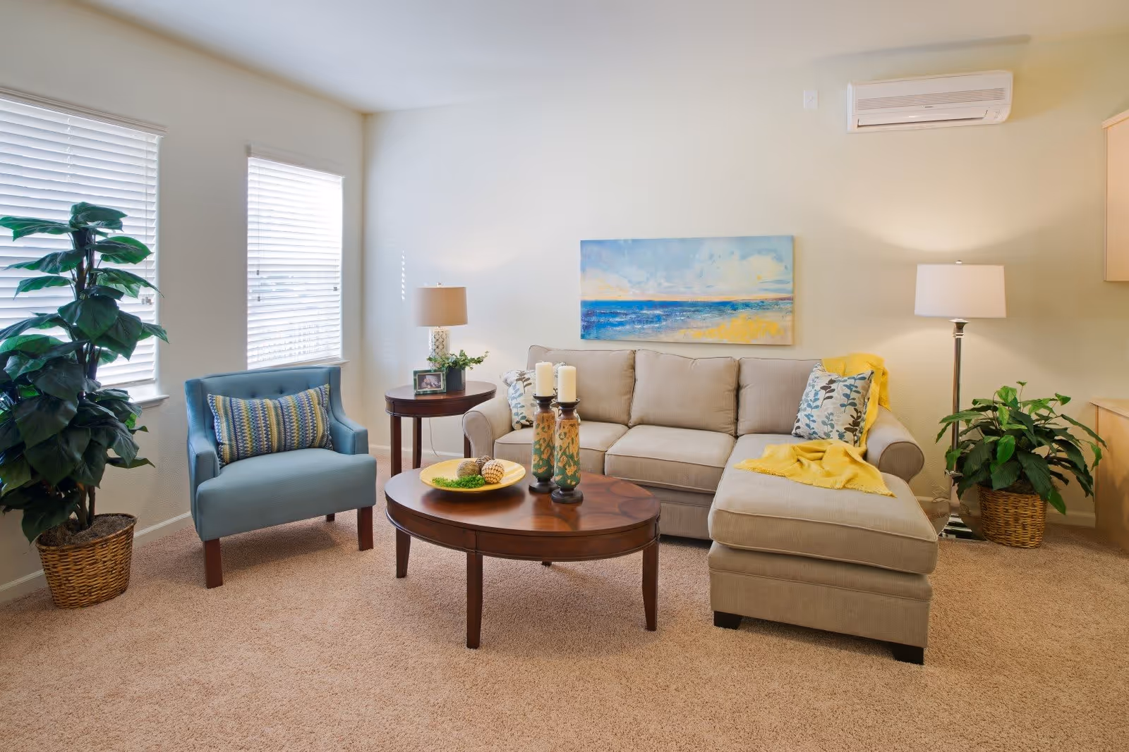 A cozy living room with a beige sectional sofa adorned with patterned pillows and a yellow throw blanket. A wooden oval coffee table with decorative items sits in front of the sofa. To the left, there is a blue armchair with a striped pillow next to a round wooden side table with a lamp and a small plant. Two large windows with white blinds let in natural light. There are two potted plants on either side of the room, and a colorful painting of a beach scene hangs on the wall above the sofa. The room has beige carpet and light-colored walls.