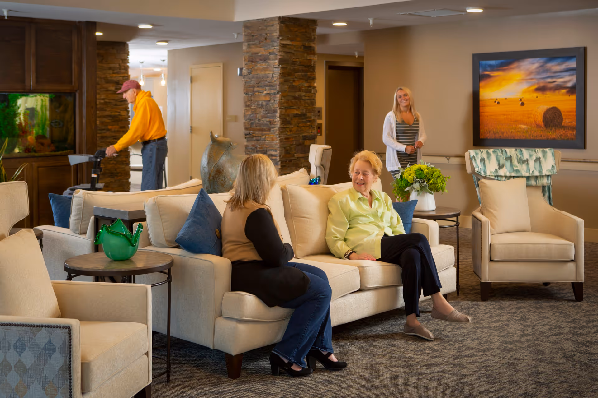 A cozy senior living facility common area with beige sofas and armchairs. Two elderly women are seated on a sofa engaged in conversation, while a younger woman stands smiling in the background. An elderly man with a walker is near a stone pillar and a large fish tank. The room is warmly lit with a framed landscape painting on the wall and a green plant on a side table.