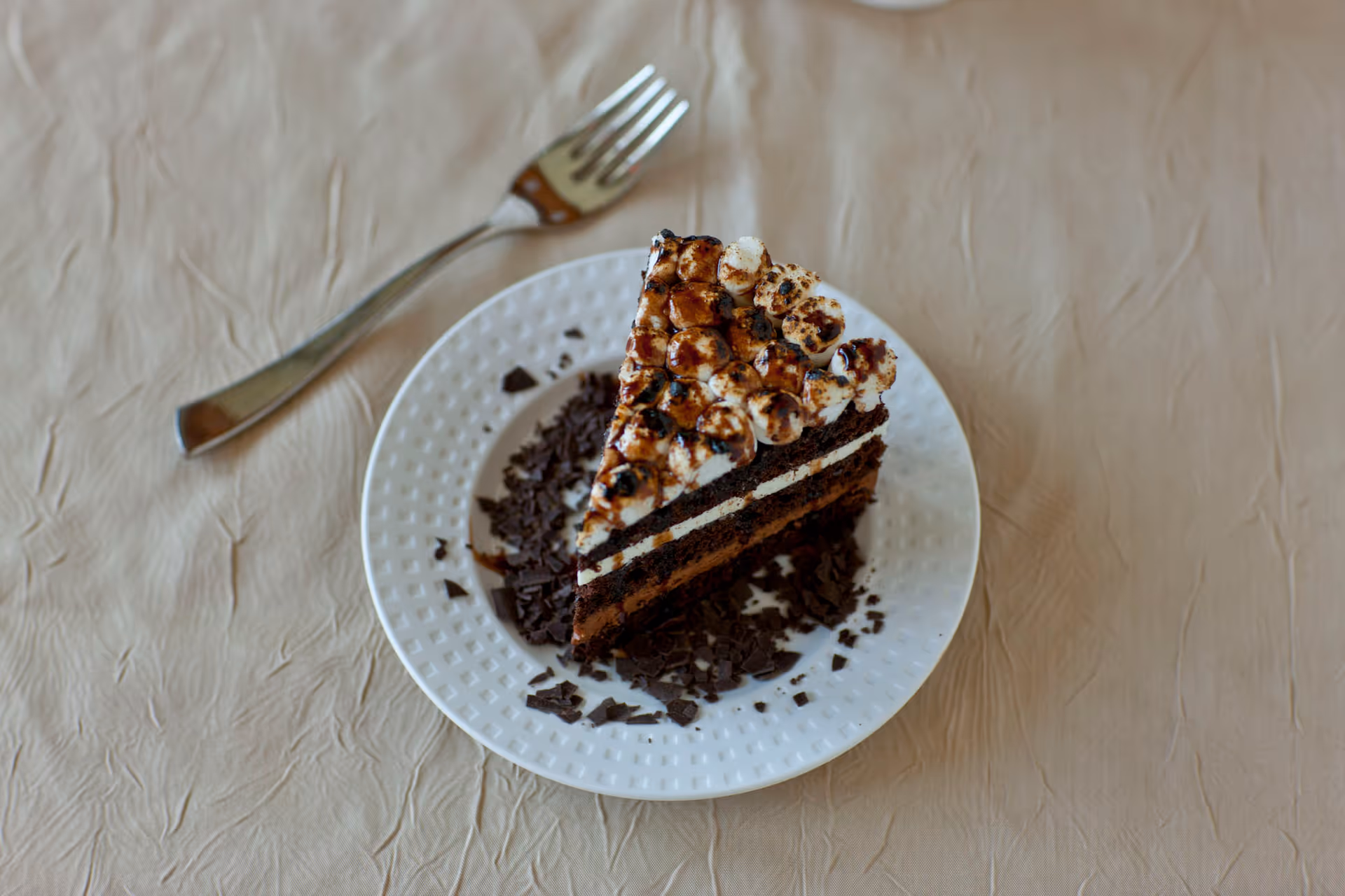 A slice of chocolate cake topped with toasted marshmallows on a white plate with chocolate shavings, placed on a beige tablecloth with a fork beside the plate.