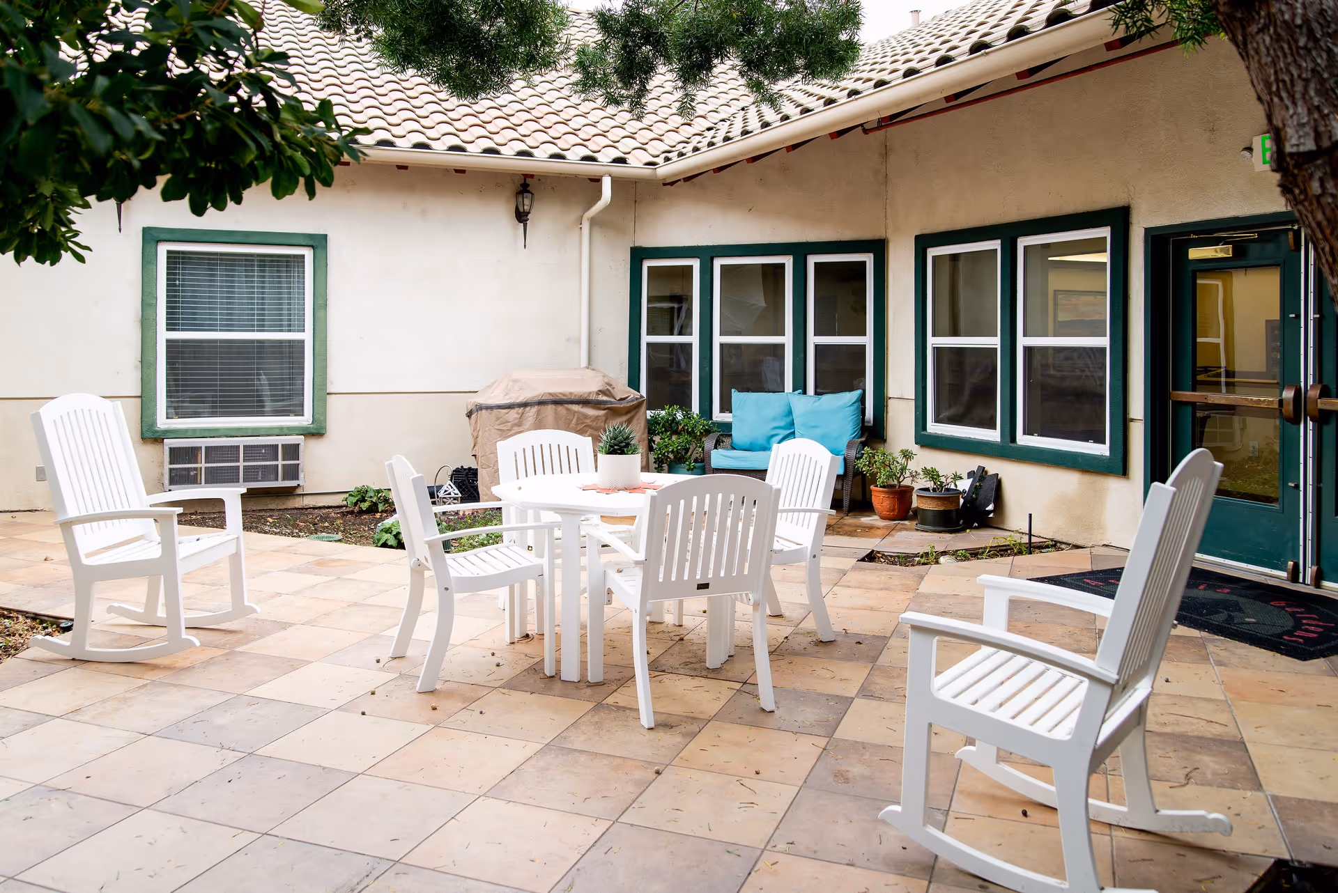 Outdoor patio area at Astoria Senior Living Tracy with white rocking chairs and a round table with chairs on a tiled floor, surrounded by windows and a door with green trim, potted plants, and a covered grill.