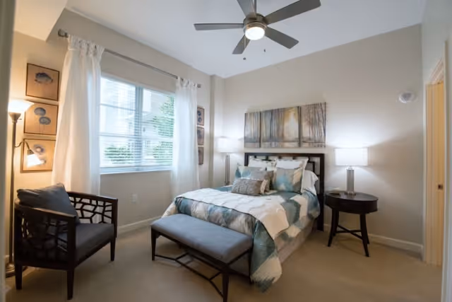 A well-lit bedroom with a bed dressed in blue and white bedding, a bench at the foot of the bed, a dark wooden chair with a cushion, two bedside tables with lamps, a ceiling fan, and a window with white curtains letting in natural light. Wall art and a floor lamp are also visible.