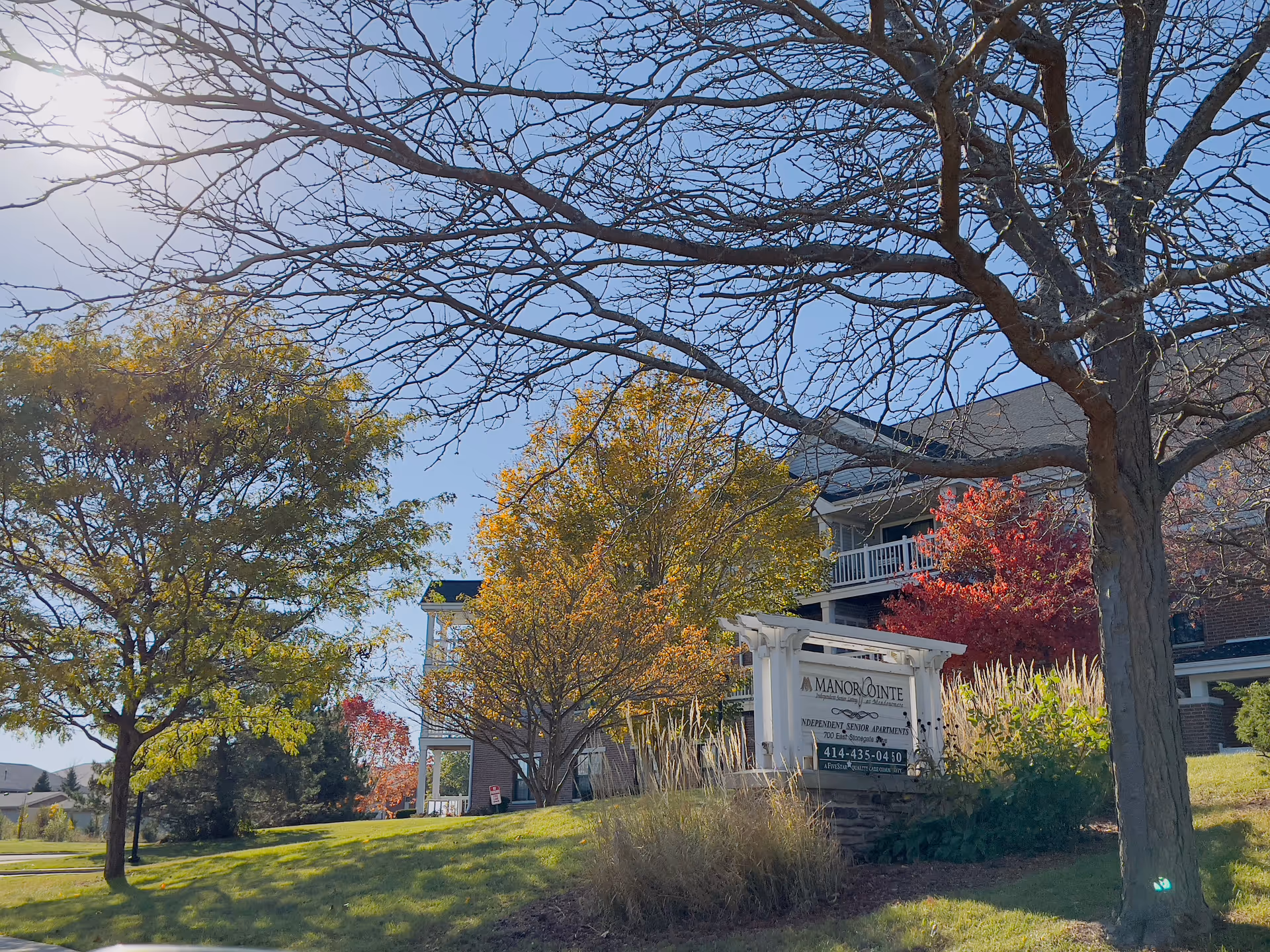 Front lawn and entrance sign for Manorpointe Apartments beside trees and a multi-story building under a clear blue sky.