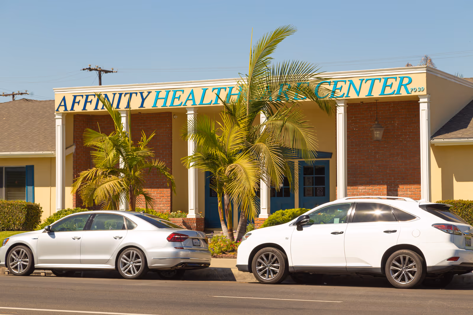 Exterior view of a building with a sign reading 'Affinity Health Care Center' partially obscured by palm trees. Two white cars are parked in front of the building on the street.