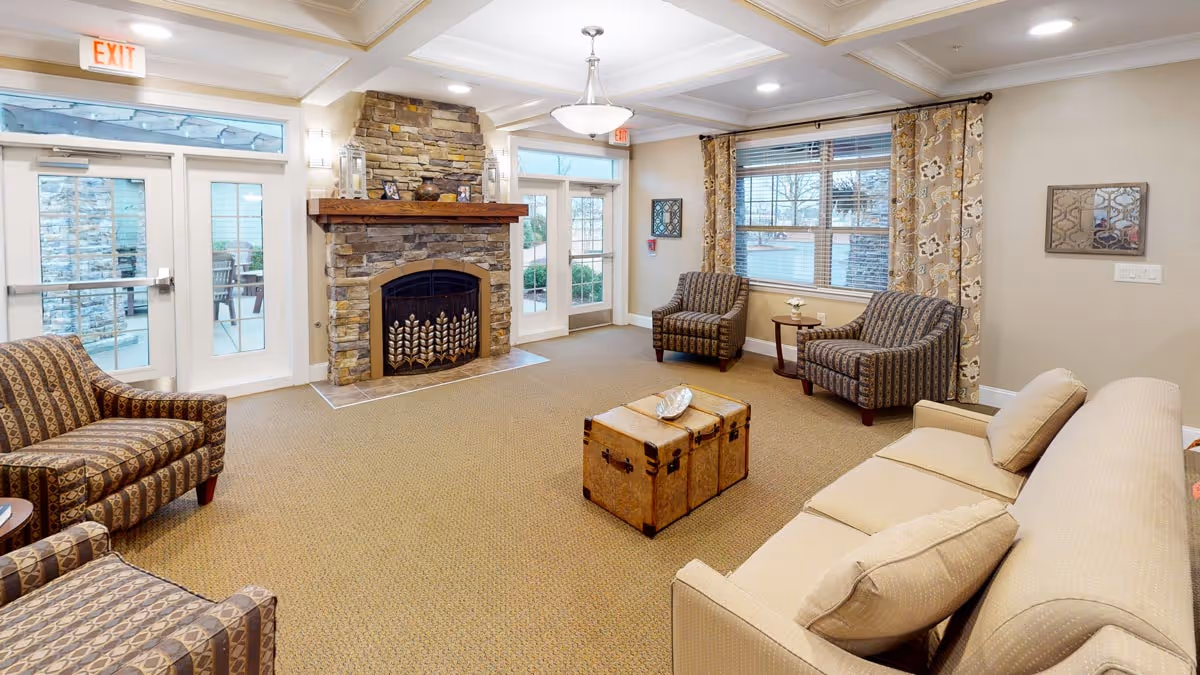 A cozy living room with a beige sofa, three patterned armchairs, a stone fireplace with decorative items on the mantel, a wooden trunk coffee table, and large windows with patterned curtains letting in natural light.