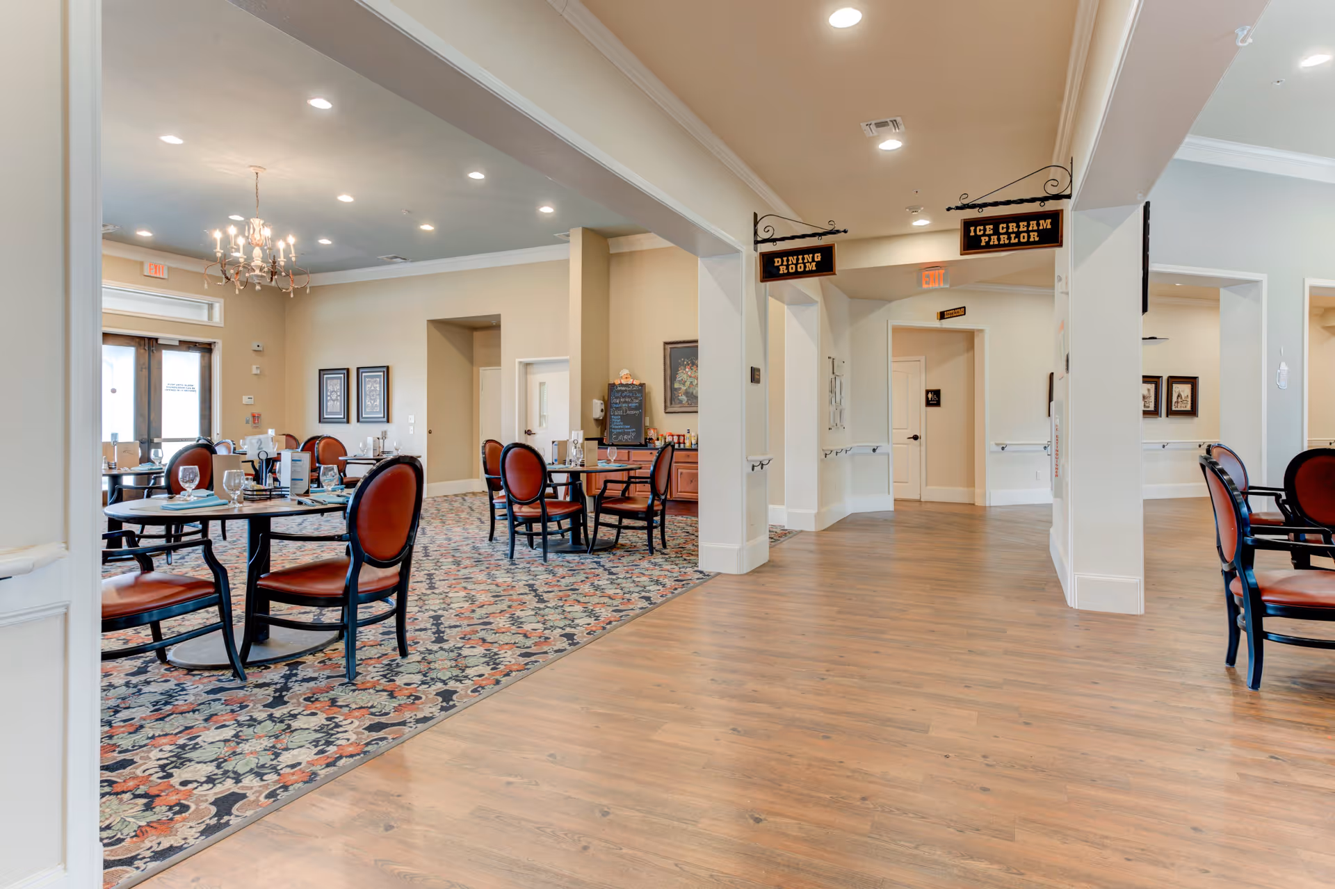 Interior view of a senior living facility dining area with round tables and red cushioned chairs on a patterned carpet. The space is well-lit with recessed lighting and a chandelier. Signs hanging from the ceiling indicate directions to the Dining Room and Ice Cream Parlor. The floor outside the carpeted area is wooden.