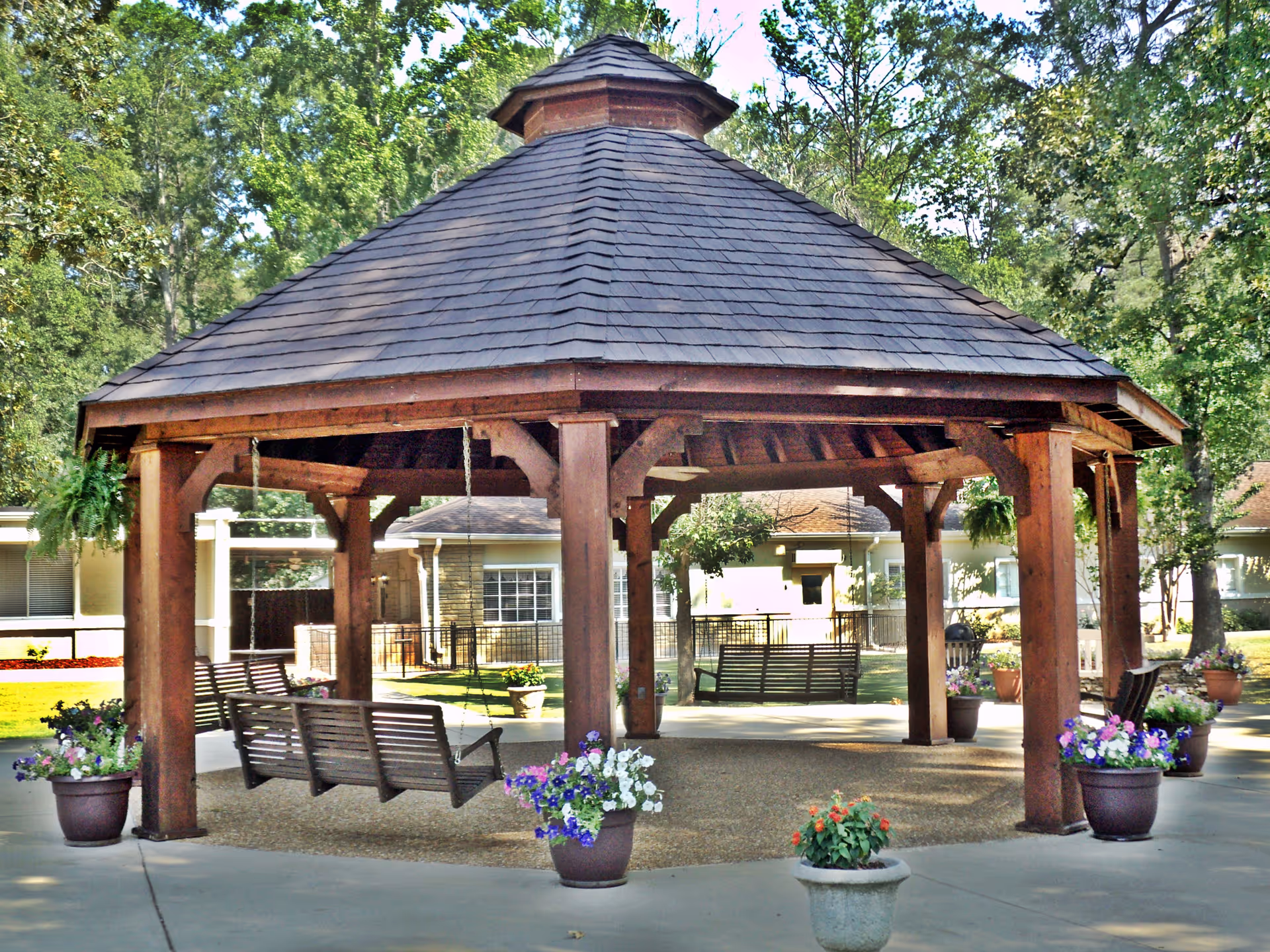 A wooden gazebo with a shingled roof located outdoors in a garden area. The gazebo has multiple wooden swings hanging from its beams and is surrounded by potted flowers and plants. Trees and a building are visible in the background.