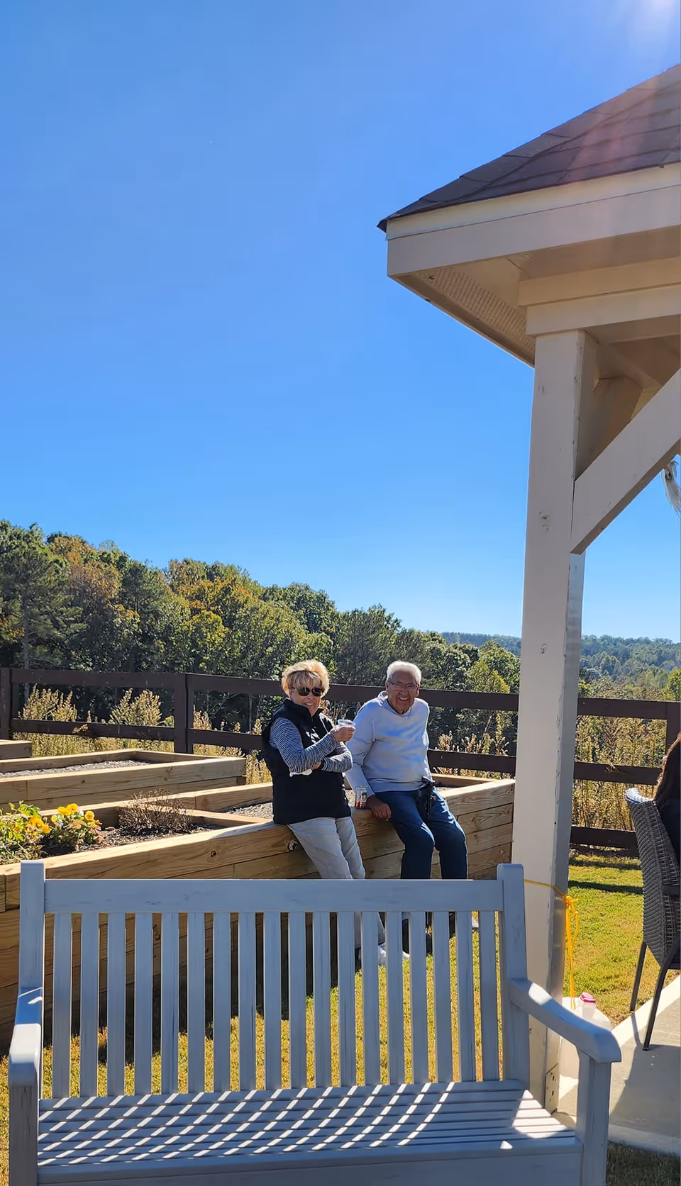 Two elderly people sitting on a raised wooden garden bed outside on a sunny day, with trees and a clear blue sky in the background. A white wooden bench is in the foreground, and part of a covered patio structure is visible on the right.