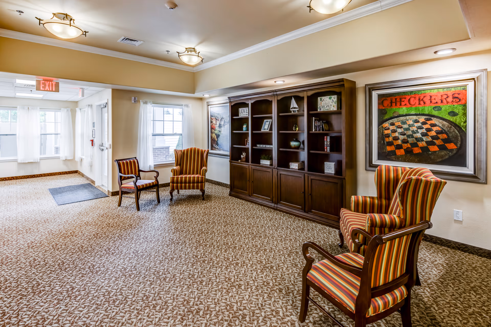 Well-lit seating area in a memory care facility with striped armchairs, a large wooden bookshelf, and a framed 'CHECKERS' painting.