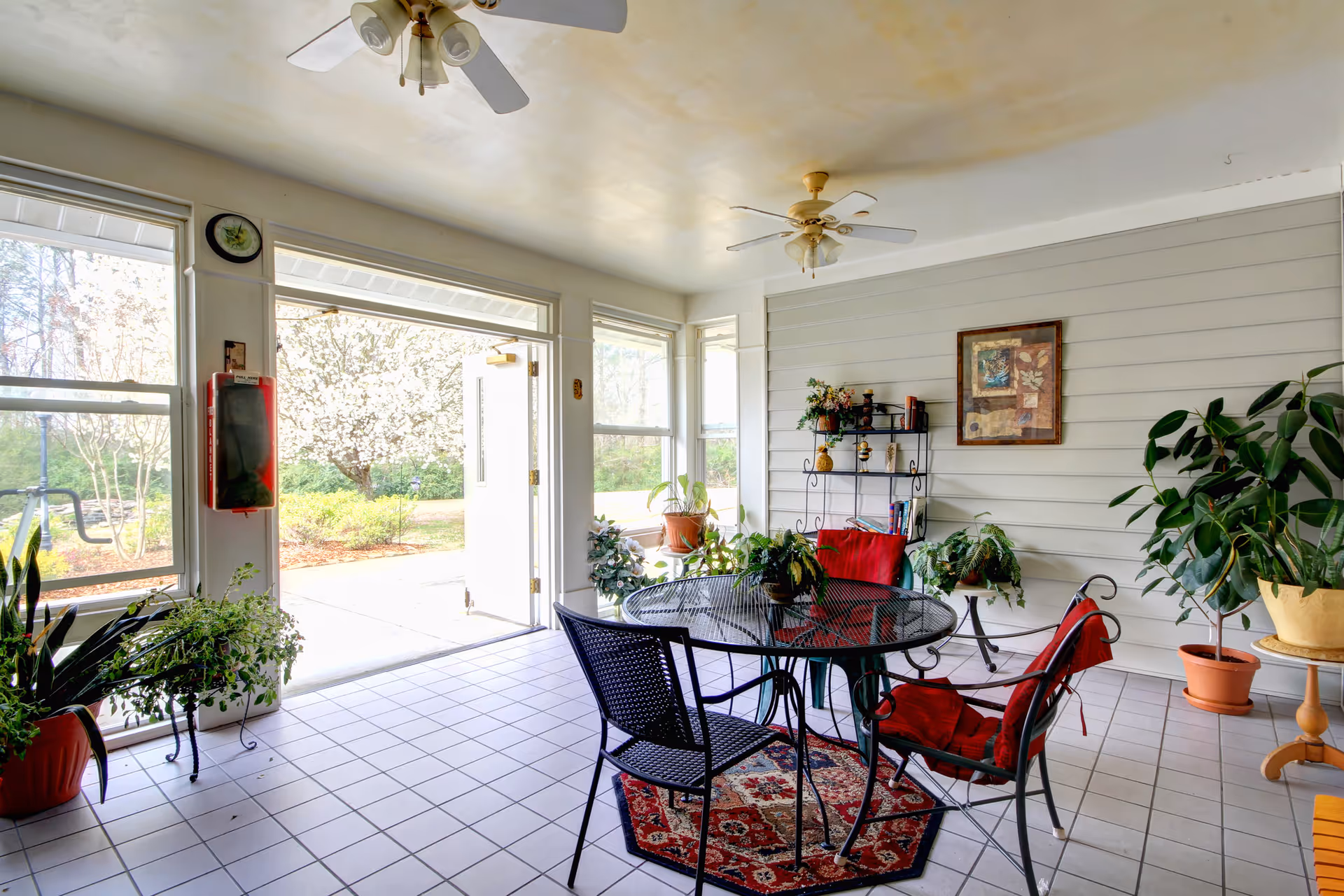A bright indoor seating area with a round black metal table and four chairs with red cushions. The room has large windows and a door open to a garden outside. Several potted plants are placed around the room, and a small shelf with decorative items and a framed picture hang on the wall. The floor is tiled, and two ceiling fans with lights are mounted on the ceiling.