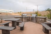 Outdoor patio area with stone picnic tables and two stainless steel barbecue grills beside a low stone wall, with a parking lot and building in the background.