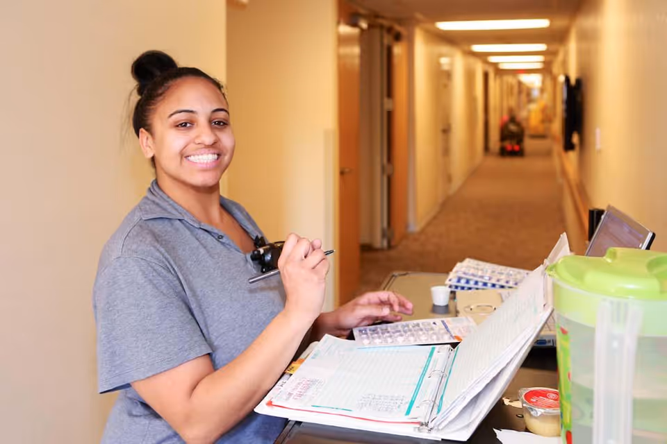 A smiling female staff member in a grey uniform stands in a hallway of a senior living facility, holding a pen and a clipboard with papers and medication blister packs. The hallway is lined with doors and has a carpeted floor, with another person visible in the distance using a mobility scooter.