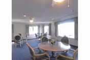 A bright common area with round tables and cushioned chairs arranged on a blue carpet. Large windows with curtains allow natural light to fill the room. A small floral centerpiece is on the nearest table.