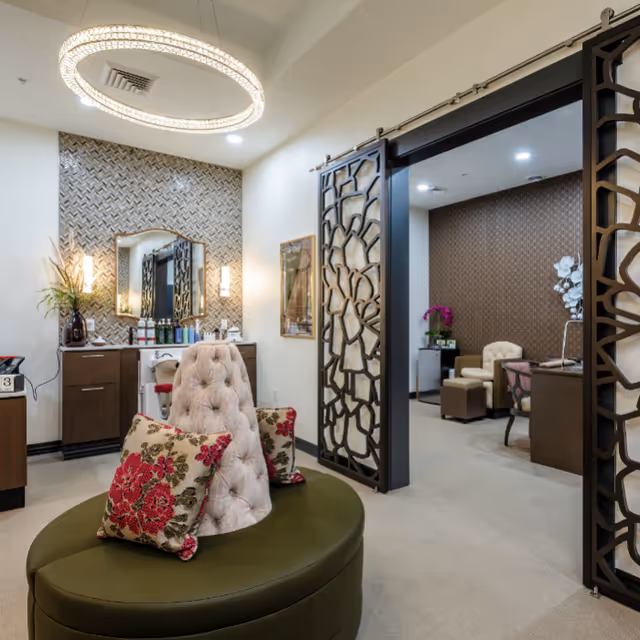 Interior view of a stylish salon or spa area in a retirement community featuring a circular olive green cushioned bench with floral pillows, a tufted salon chair, a vanity with a mirror and hair care products, decorative wall panels, and modern lighting fixtures.