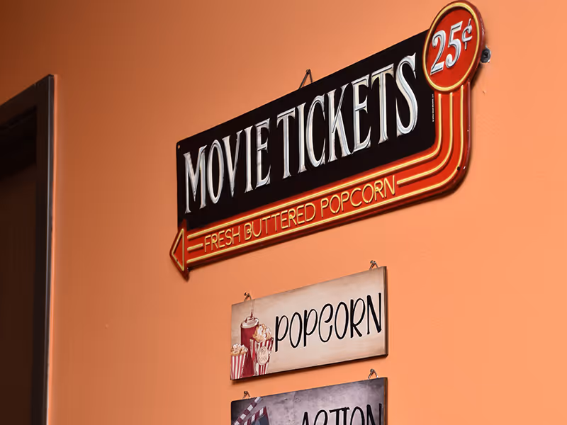 A wall painted in orange with three hanging signs. The top sign is shaped like an arrow and reads 'MOVIE TICKETS 25¢ FRESH BUTTERED POPCORN' in white and red letters. Below it, there is a smaller rectangular sign with an illustration of popcorn and a drink, labeled 'POPCORN'.