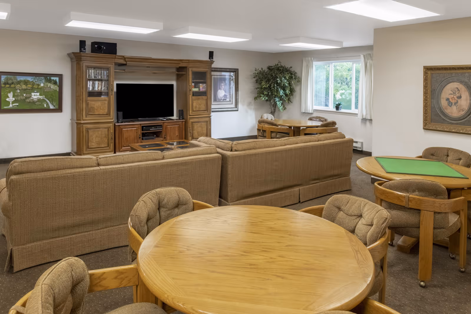 A cozy common area with brown cushioned sofas facing a wooden entertainment center with a TV. There are round wooden tables with padded chairs around the room, a large window with white curtains, framed artwork on the walls, and a potted plant in the corner.