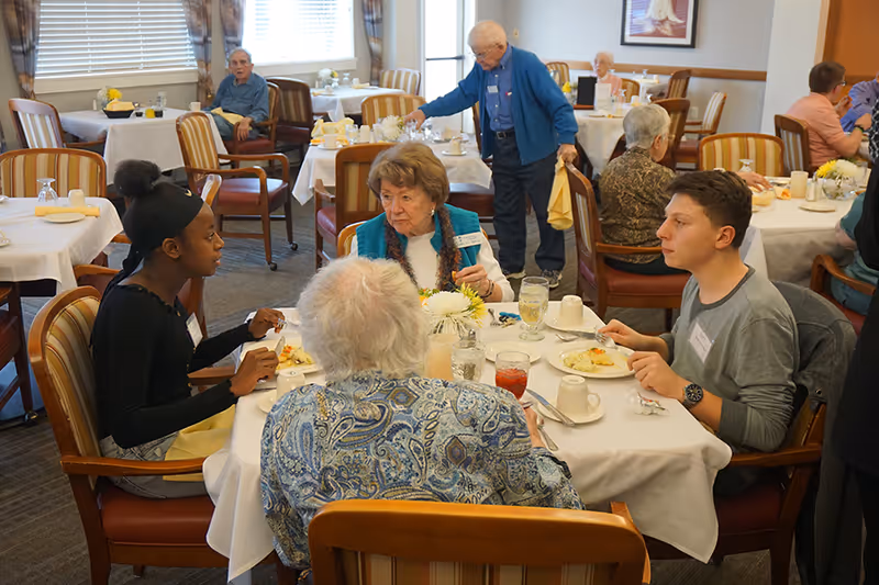 A group of elderly people and a young woman are seated around a dining table in a senior living facility dining room. They are engaged in conversation while having a meal. Other residents are visible in the background, seated at tables with white tablecloths and floral centerpieces. The room has large windows with curtains and wooden chairs with striped cushions.
