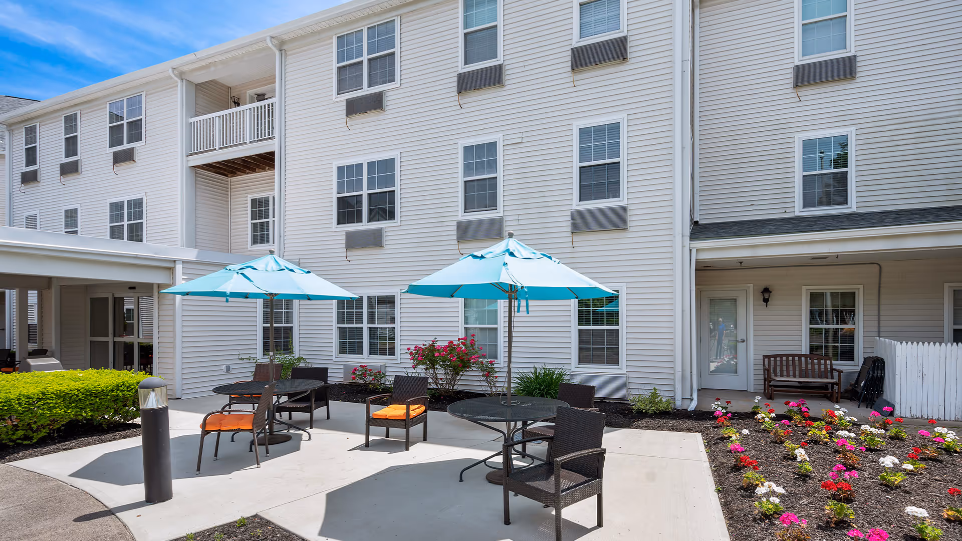 Outdoor patio area at Holiday Lodge at Cold Spring with round tables, chairs, and blue umbrellas. The patio is surrounded by flower beds with colorful flowers and a three-story white building with multiple windows in the background under a blue sky.