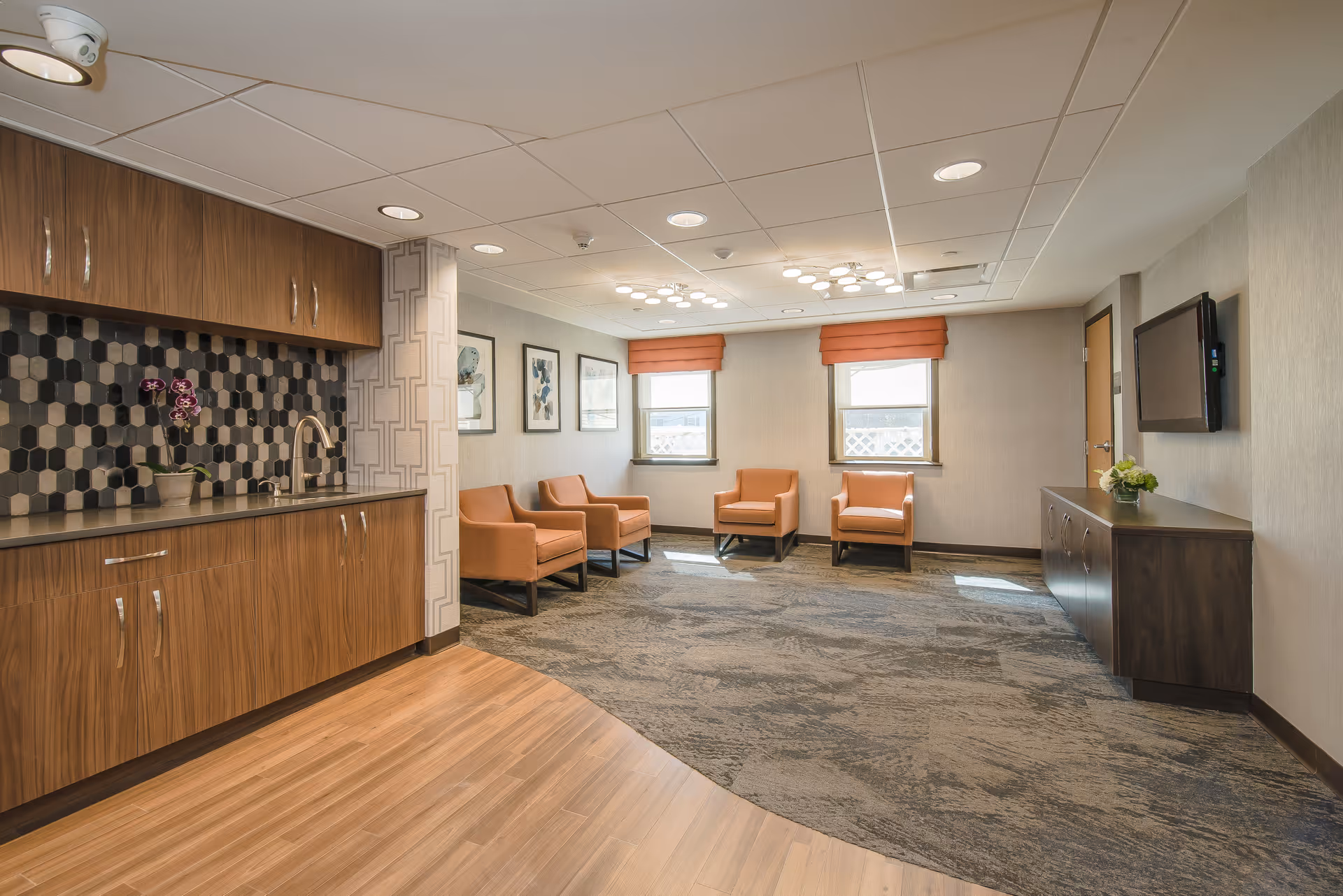 A modern interior room with a kitchenette on the left featuring wooden cabinets, a sink, and a hexagonal tile backsplash. The room has four orange armchairs arranged near two windows with orange blinds. On the right wall, there is a mounted flat-screen TV above a dark wooden cabinet with a small plant on top. The floor transitions from wood near the kitchenette to carpet in the seating area.