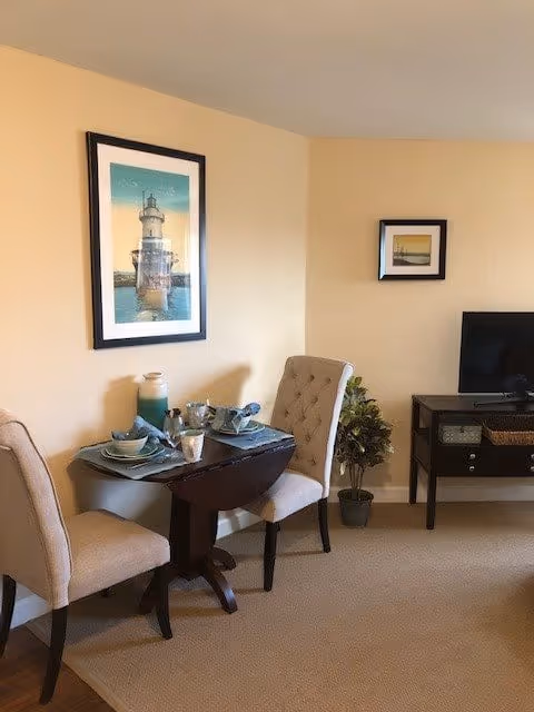A small dining area with a dark wooden table set for two with plates, bowls, cups, and a pitcher. Two beige upholstered chairs are placed on either side of the table. On the wall above the table is a framed picture of a lighthouse. To the right, there is a small black TV stand with a flat-screen TV on top, a basket on the lower shelf, and a potted plant beside it. The walls are painted light beige and the floor is carpeted.