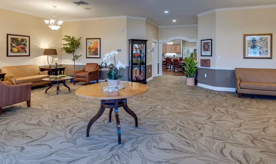 A spacious senior living facility common area with beige walls and patterned carpet. The room features several seating arrangements including sofas and armchairs in warm tones, a round wooden table with a potted orchid in the center, framed artwork on the walls, and a tall glass display cabinet. In the background, an open doorway leads to a dining area with tables and chairs.