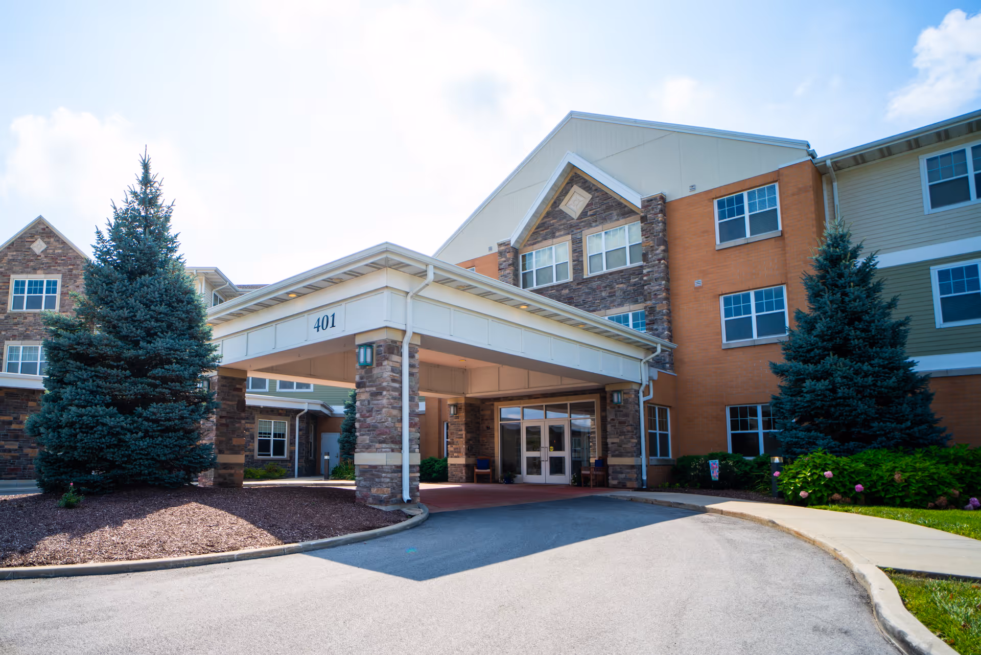 Front entrance of a three-story senior living building with a covered porte-cochere labeled 401, flanked by evergreen trees.