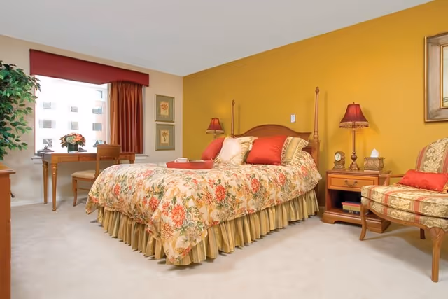 A cozy bedroom with a floral-patterned bedspread on a wooden bed frame, two red lamps on wooden nightstands, a floral upholstered chair with a red pillow, a wooden desk with a chair near a window with red curtains, and a yellow accent wall.