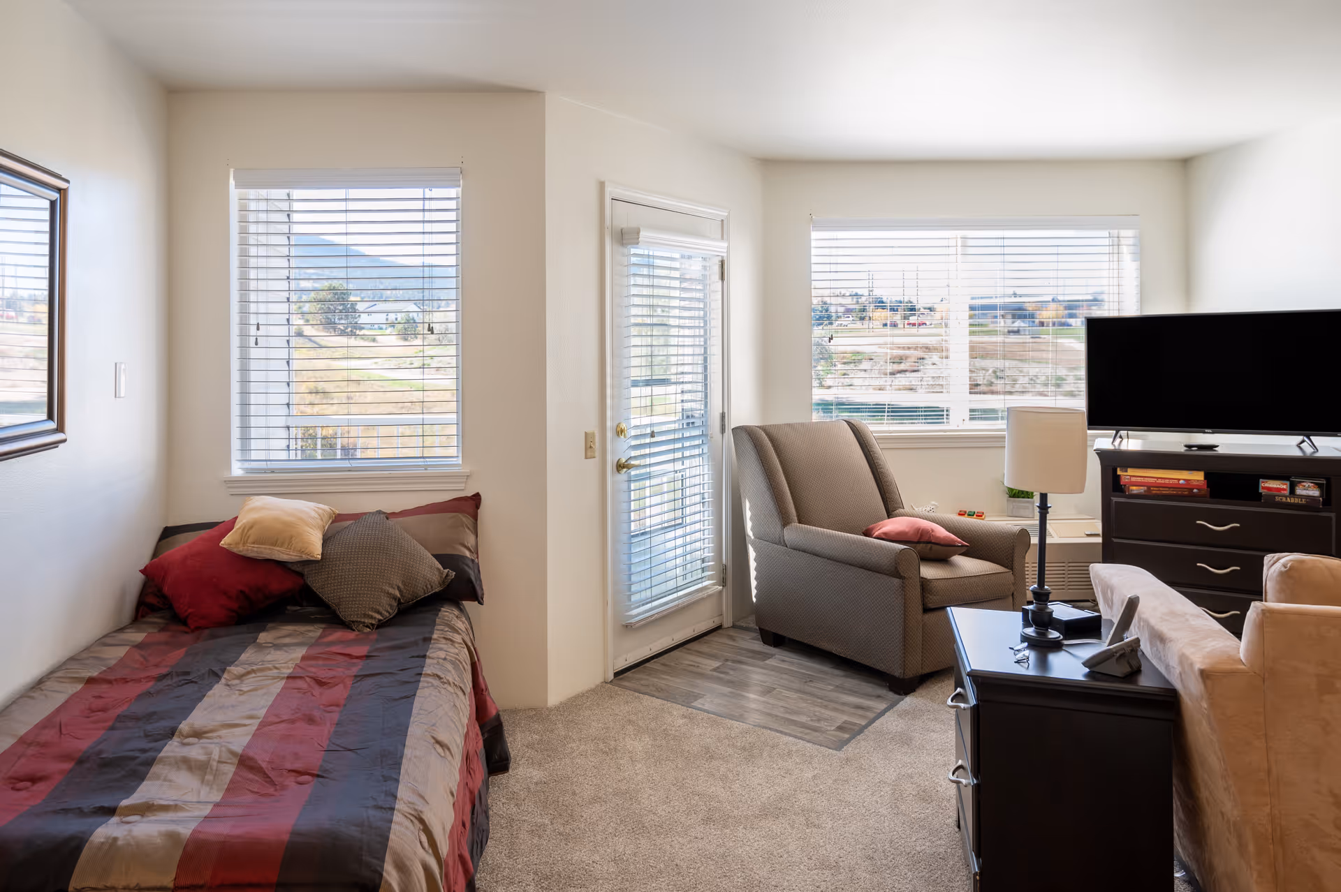 Studio apartment interior with a bed on the left, an armchair and side table near windows, and a TV on a dresser.
