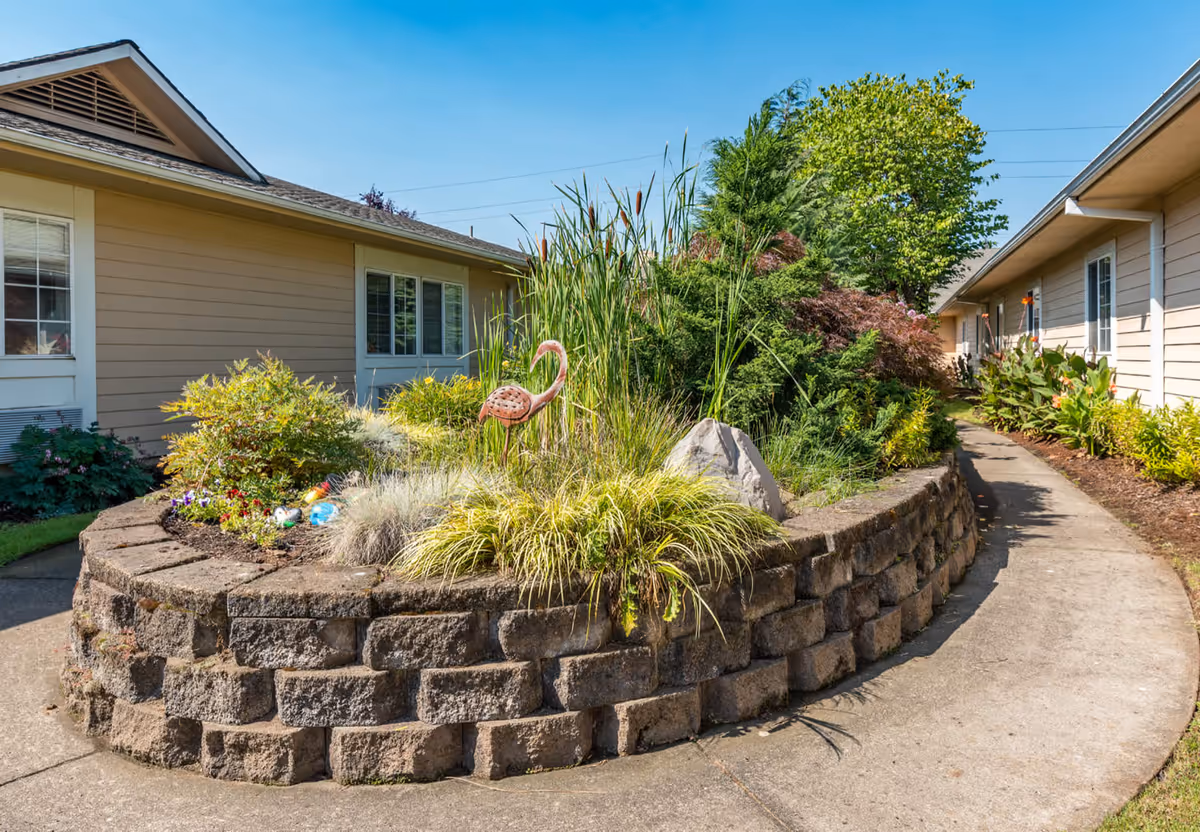 Outdoor garden area with a raised stone planter filled with various plants and a metal flamingo decoration, surrounded by beige single-story buildings under a clear blue sky.