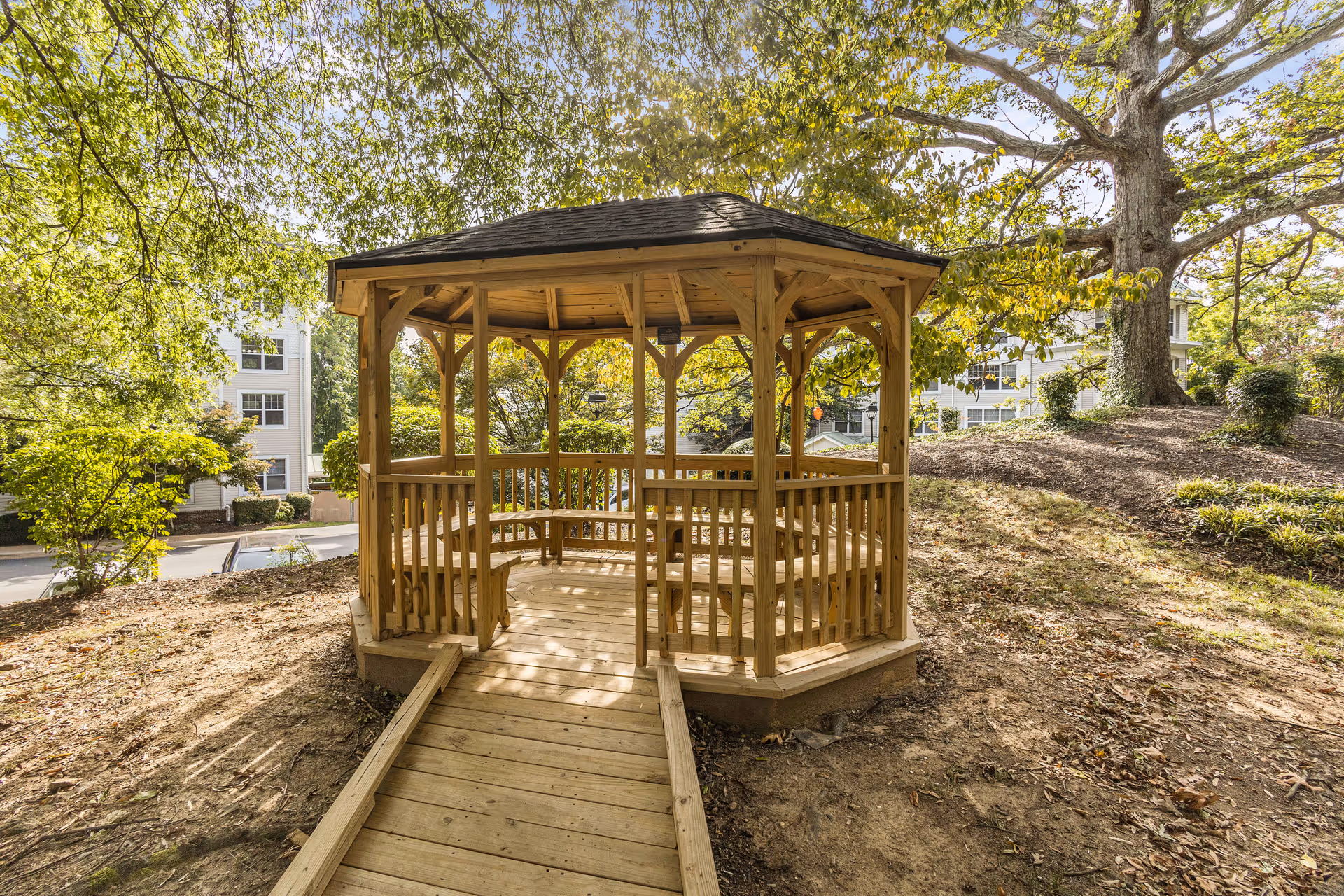 Wooden octagonal gazebo with a ramp and built-in benches amid trees on a grassy slope with buildings visible behind.