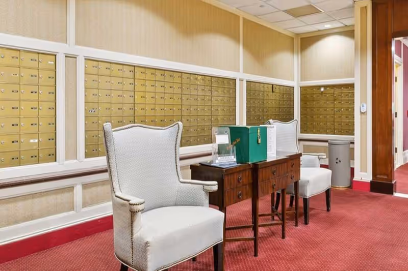 Interior view of a mailroom area with multiple rows of gold-colored mailboxes mounted on the walls. In front of the mailboxes, there are two white upholstered armchairs and a wooden table with a green box and some papers on it. The floor is carpeted in red, and the walls are beige with white trim.
