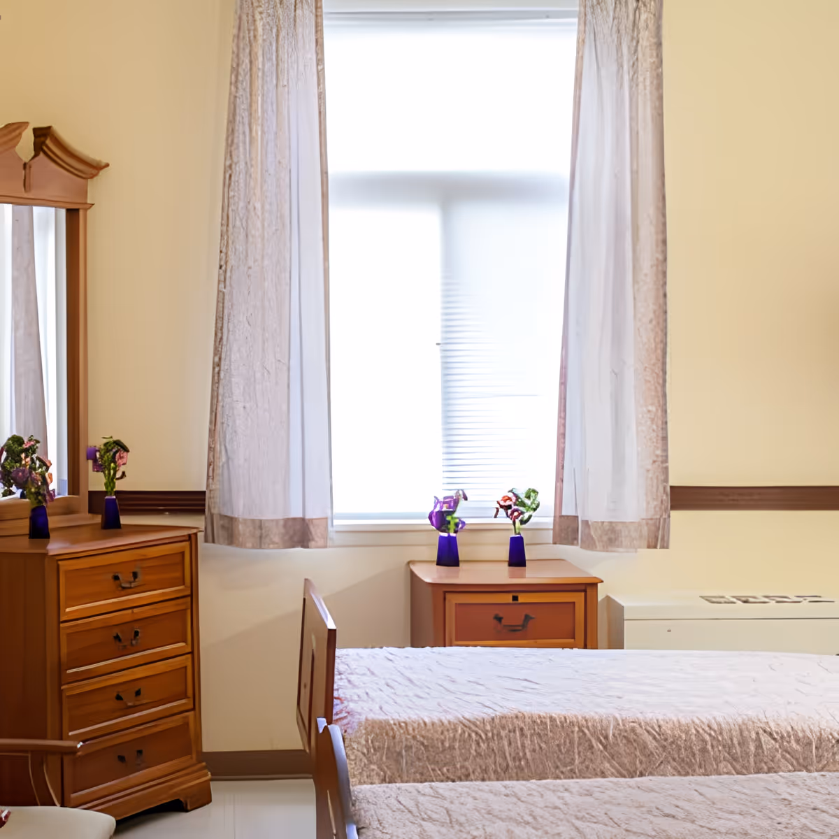 A bedroom with two single beds covered in beige bedspreads, a wooden dresser with a mirror, a wooden nightstand with two small vases holding flowers, and a window with sheer curtains allowing light to enter the room.