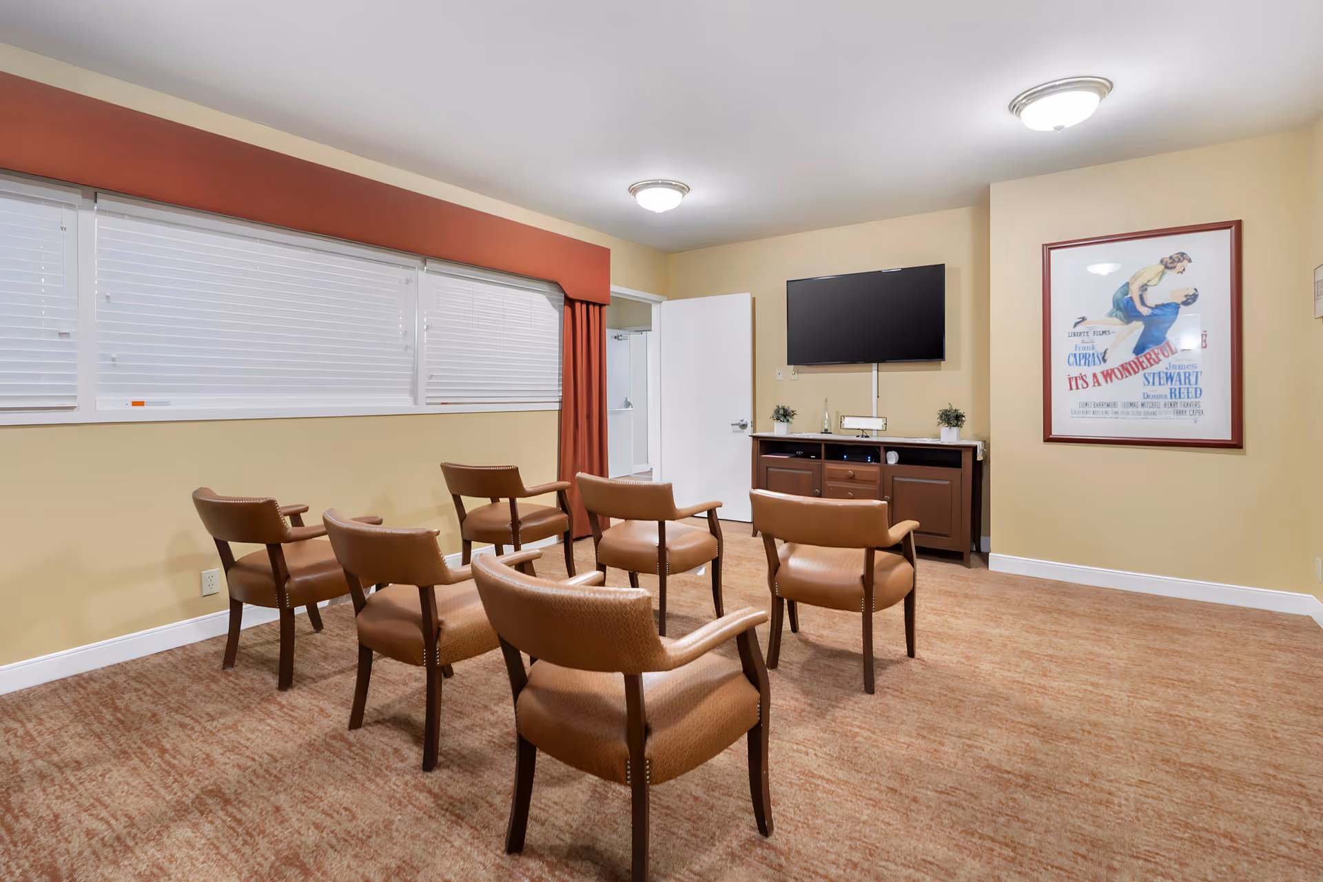A small meeting or media room with six brown leather chairs arranged in two rows facing a wall-mounted flat screen TV. Below the TV is a wooden cabinet with decorative plants and a bottle. The room has beige walls, a large window with white blinds and a red valance, and a framed vintage movie poster on the wall. The door to the room is open.