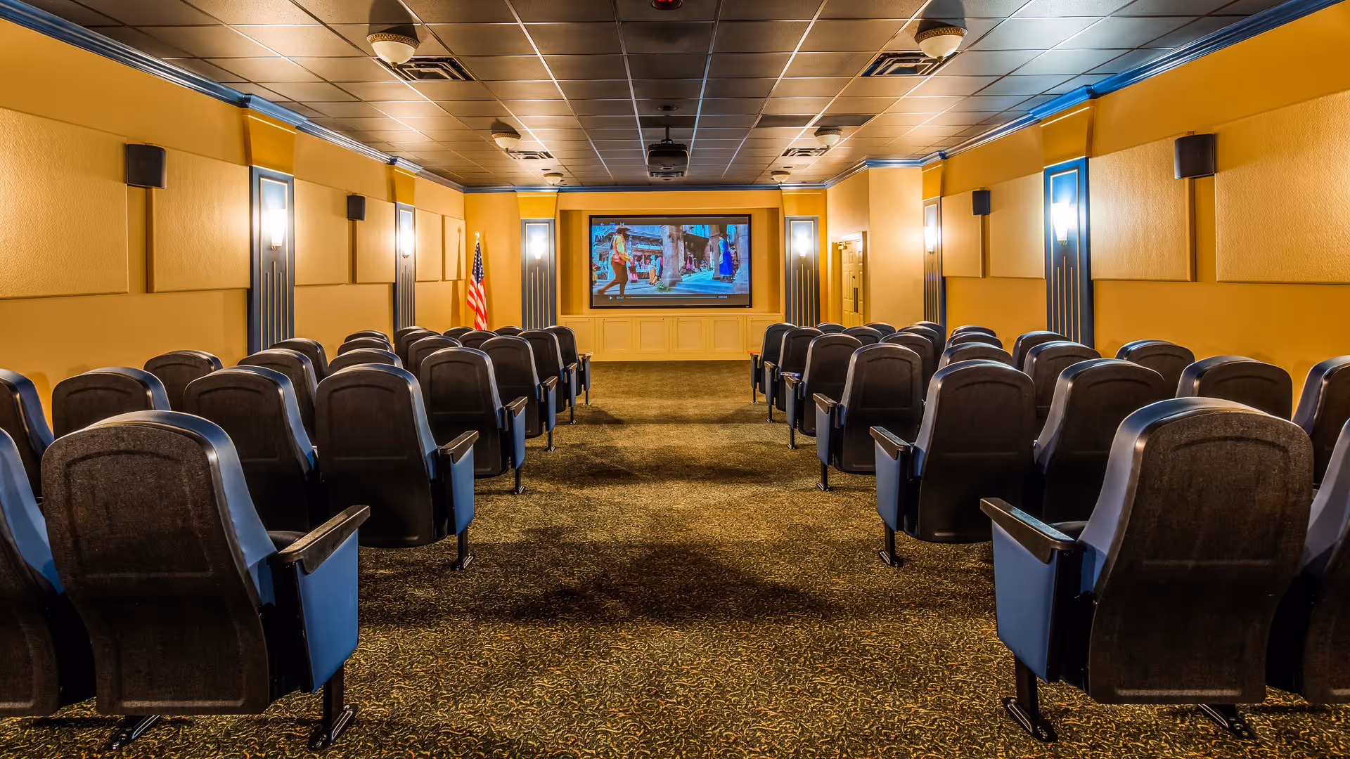 A small theater room with rows of black cushioned seats facing a large screen at the front. The walls are painted yellow with decorative panels and wall-mounted lights. An American flag is positioned to the left of the screen.