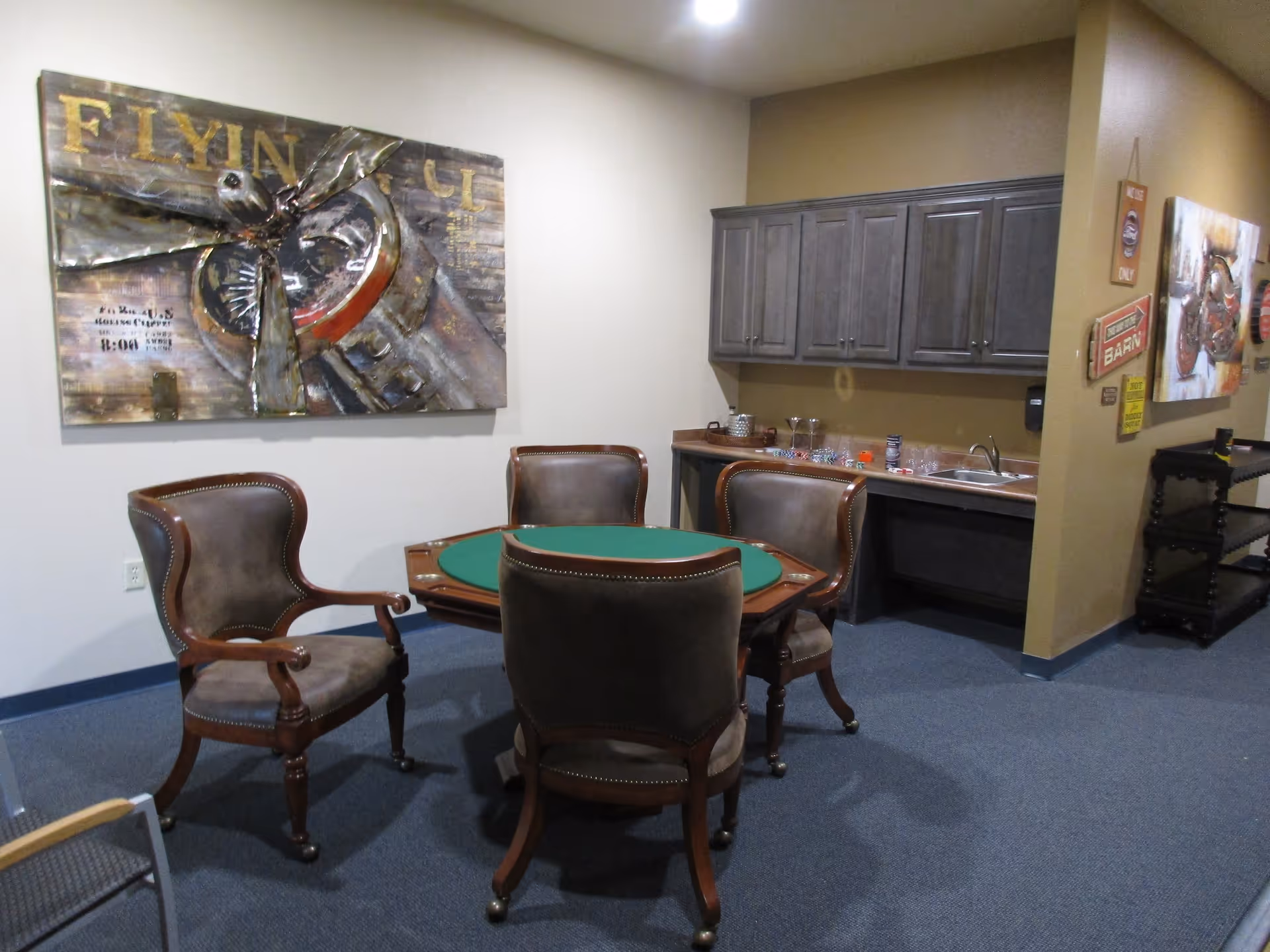 A room with a round green felt card table surrounded by four brown leather chairs with wooden legs. On the right side, there is a countertop with a sink and upper cabinets. The walls are decorated with metal and painted artwork, including a large piece featuring an airplane propeller. The floor is carpeted in blue.
