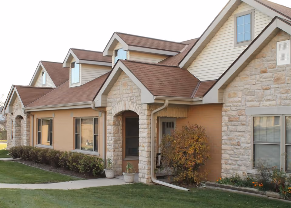 Front exterior of a low-rise stone-and-siding building with arched entryways, dormer windows, and a small lawn with shrubs.