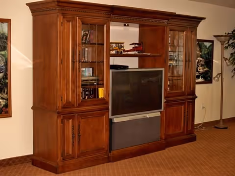A large wooden entertainment center with glass-paneled cabinets on either side, shelves with decorative items including model airplanes, and a television in the center. The unit is placed against a beige wall with framed artwork and a tall floor lamp nearby.