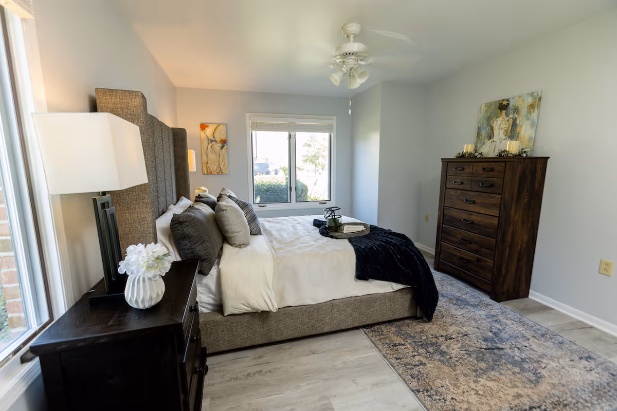 A bright and neatly arranged bedroom with a large upholstered bed featuring multiple pillows and a dark throw blanket. There is a dark wooden nightstand with a white vase holding flowers and a modern lamp. A window with blinds lets in natural light. Across from the bed is a tall dark wooden dresser with decorative items and a painting above it. The room has light-colored walls and flooring with a patterned area rug.