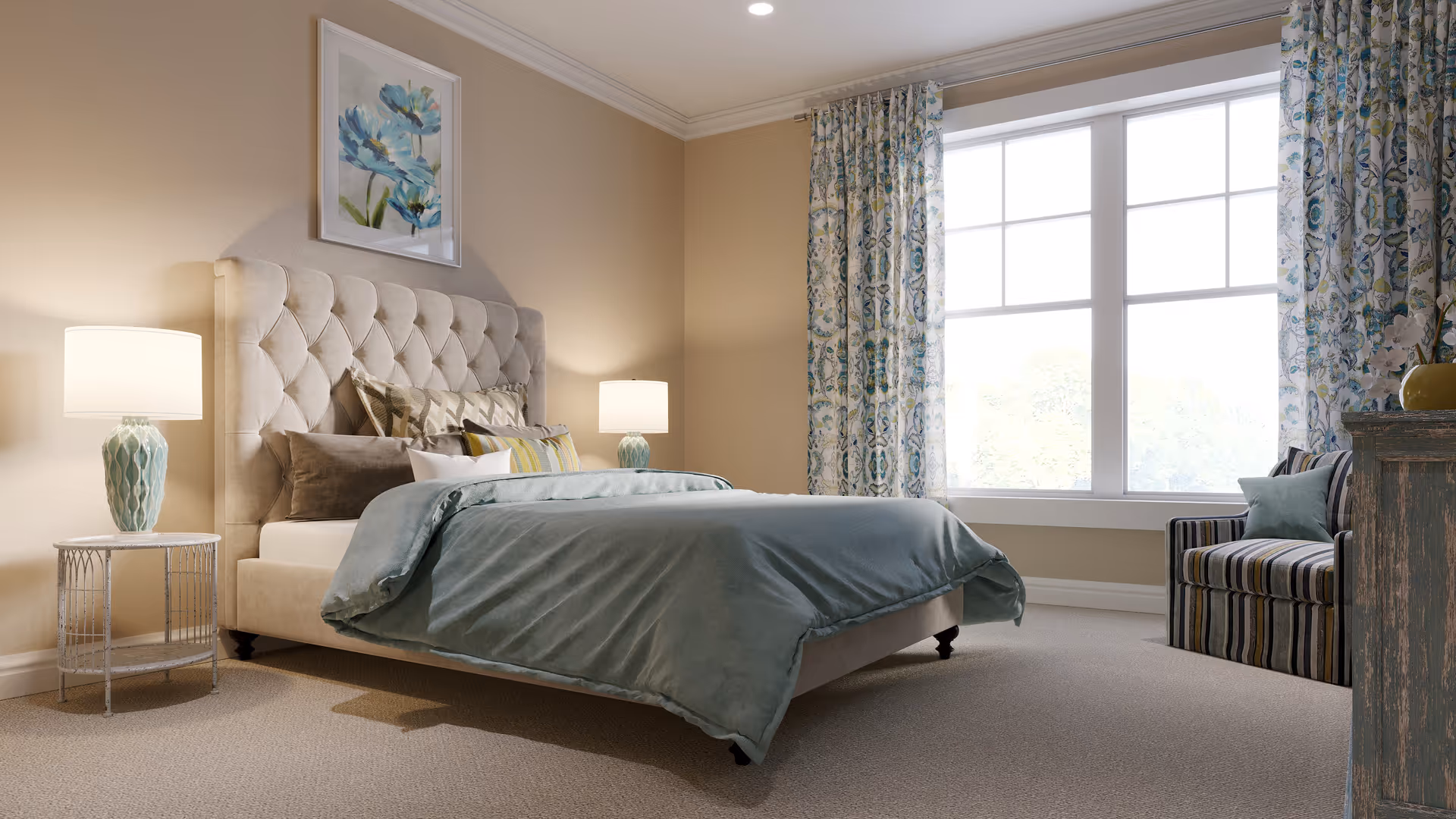 A cozy bedroom with a large tufted beige headboard bed covered with a light blue comforter and multiple pillows. There are two bedside tables with matching lamps, a large window with patterned curtains, a striped armchair, and a framed floral artwork on the wall.
