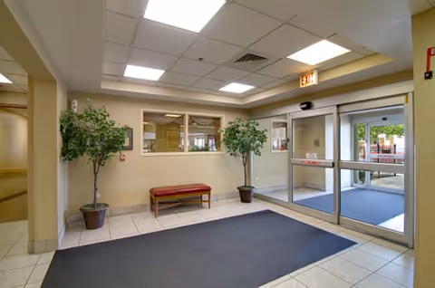 Entrance area of a senior living facility with glass automatic sliding doors, a large dark floor mat, two potted plants, a red bench, and a window looking into an interior room.