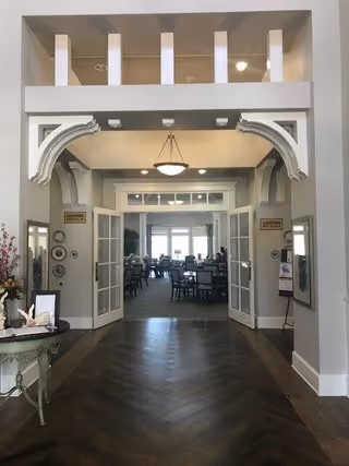 View through an entrance hallway with decorative white archways leading into a dining area with multiple tables and chairs. The dining room has large windows at the far end letting in natural light. The hallway has dark wood flooring and light gray walls with some framed decorations and signs on either side.