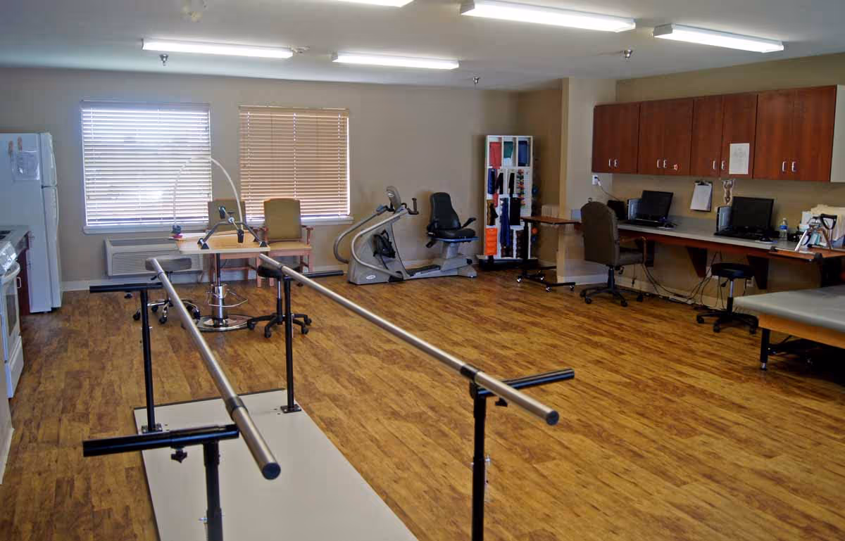 A spacious rehabilitation room with parallel bars, exercise machines, desks with computers, and wood-look flooring.
