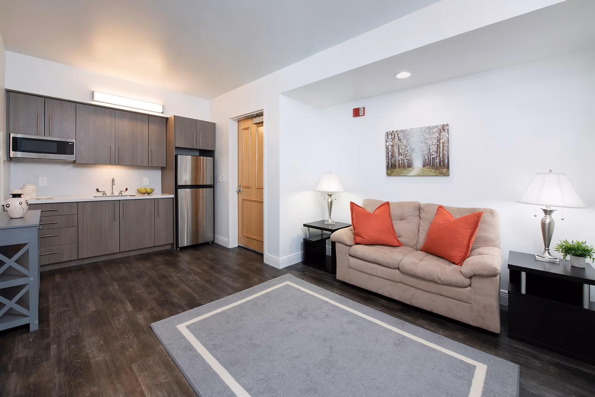 A modern living space featuring a small kitchen area with gray cabinets, a microwave, and a stainless steel refrigerator on the left. To the right, there is a beige couch with two red throw pillows, flanked by two black side tables each with a lamp. A painting of a forest path hangs on the white wall above the couch. The floor is dark wood with a gray area rug in front of the couch.
