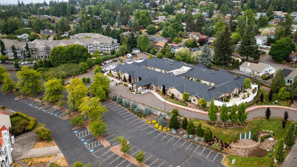 Aerial view of Sunnyside Meadows Memory Care facility surrounded by trees and greenery, with a large parking lot in the foreground and residential neighborhood in the background.