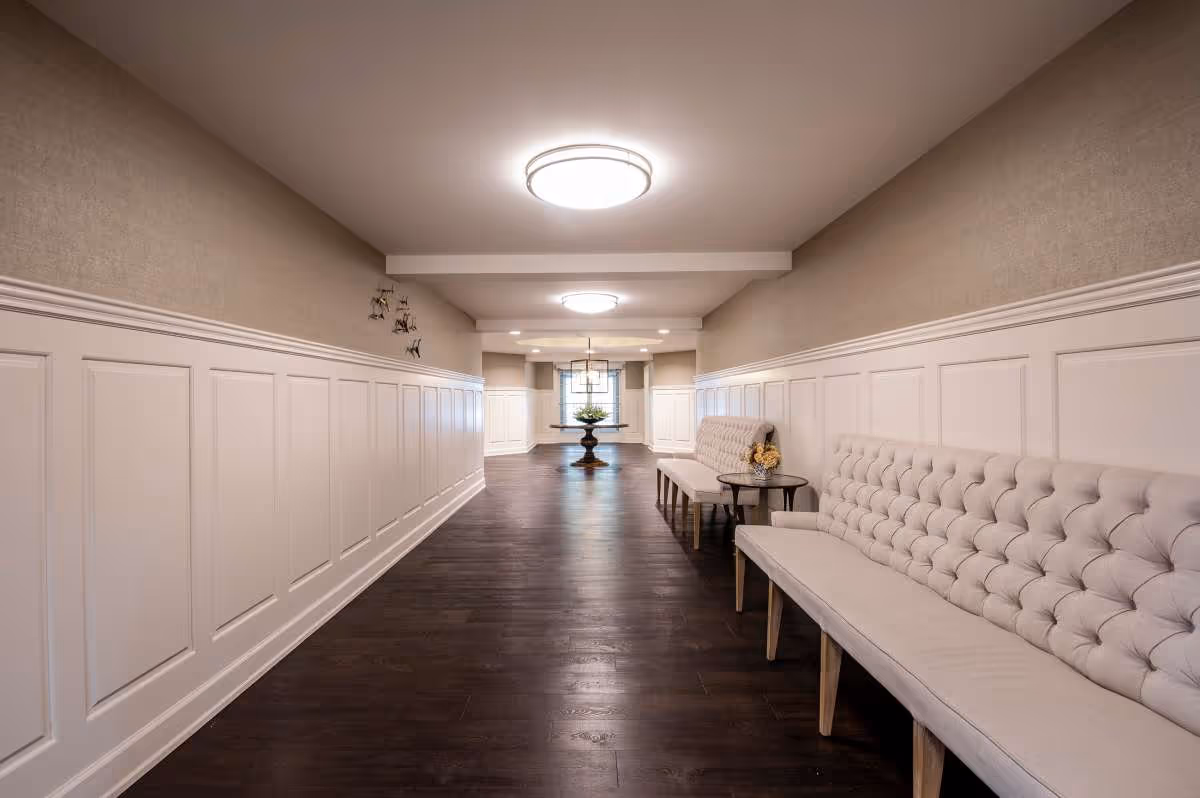 A long, well-lit hallway with dark wooden floors and beige walls featuring white wainscoting. On the right side, there are two light gray tufted benches with a small round table between them holding a small floral arrangement. At the end of the hallway, there is a round table with a decorative plant and a window letting in natural light.