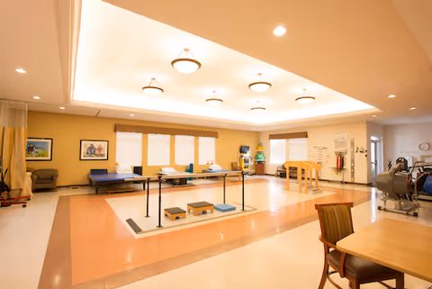 A spacious physical therapy or rehabilitation room with exercise equipment including parallel bars, steps, and therapy beds. The room has warm yellow walls, large windows with blinds, ceiling lights, and a few chairs and tables.
