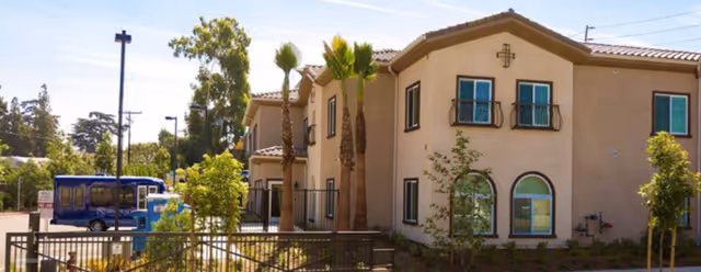 Two-story beige stucco memory care building with palm trees, a fenced entrance and a parked blue shuttle van.