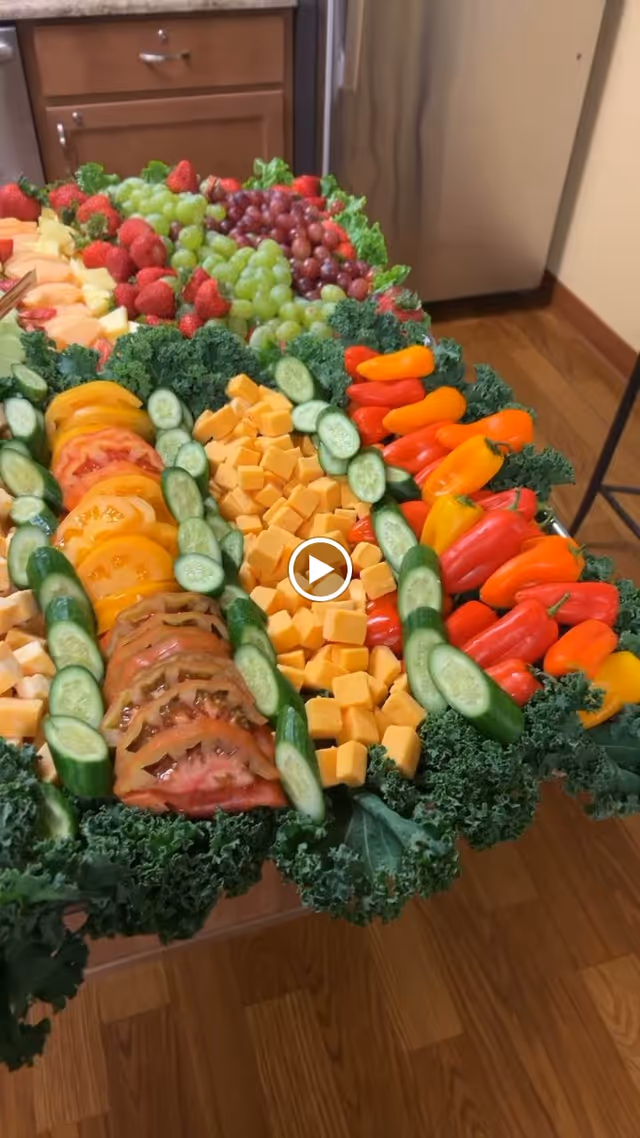 A colorful platter of fresh fruits, vegetables, and cheese cubes arranged on a bed of kale. The platter includes green and red grapes, strawberries, sliced cucumbers, yellow and red mini bell peppers, sliced tomatoes, and cubed cheddar cheese. The platter is set on a wooden floor with kitchen cabinets and a refrigerator in the background.