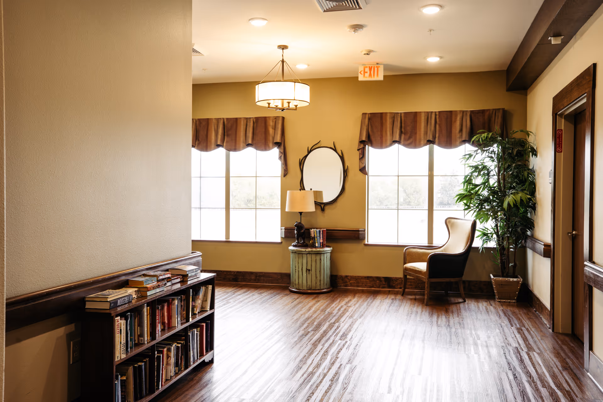 A cozy interior corner of a senior living facility with wooden flooring, a small bookshelf filled with books, a round side table with a lamp and books, a decorative mirror on the wall, two large windows with brown valances, a comfortable armchair, and a potted plant near a closed door.
