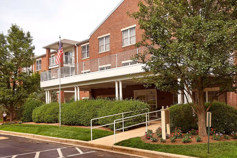 Exterior view of a red brick senior living facility named Bell Trace with a covered entrance, an American flag on a flagpole, well-maintained bushes, trees, and flower beds along the sidewalk and parking area.