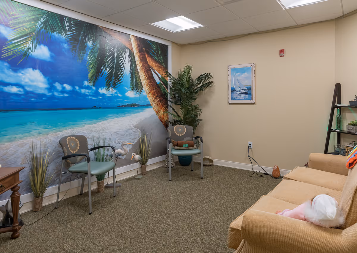 A cozy room with a large tropical beach mural on one wall, two chairs with armrests, a beige couch with a doll sitting on it, a potted plant, a small wooden table, a framed picture of a sailboat on the wall, and a black shelving unit with decorative items.